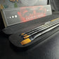 Andrew Cadima brush set in an open black case, with gold bristles and black handles beside the labeled sleeve.