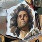 Portrait of a curly-haired man looking upward on a shaped panel, credited to Gustavo Ramos.