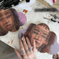 Two realistic painted faces on cloud-shaped panels beneath black-handled brushes, with an artist's hand in front.