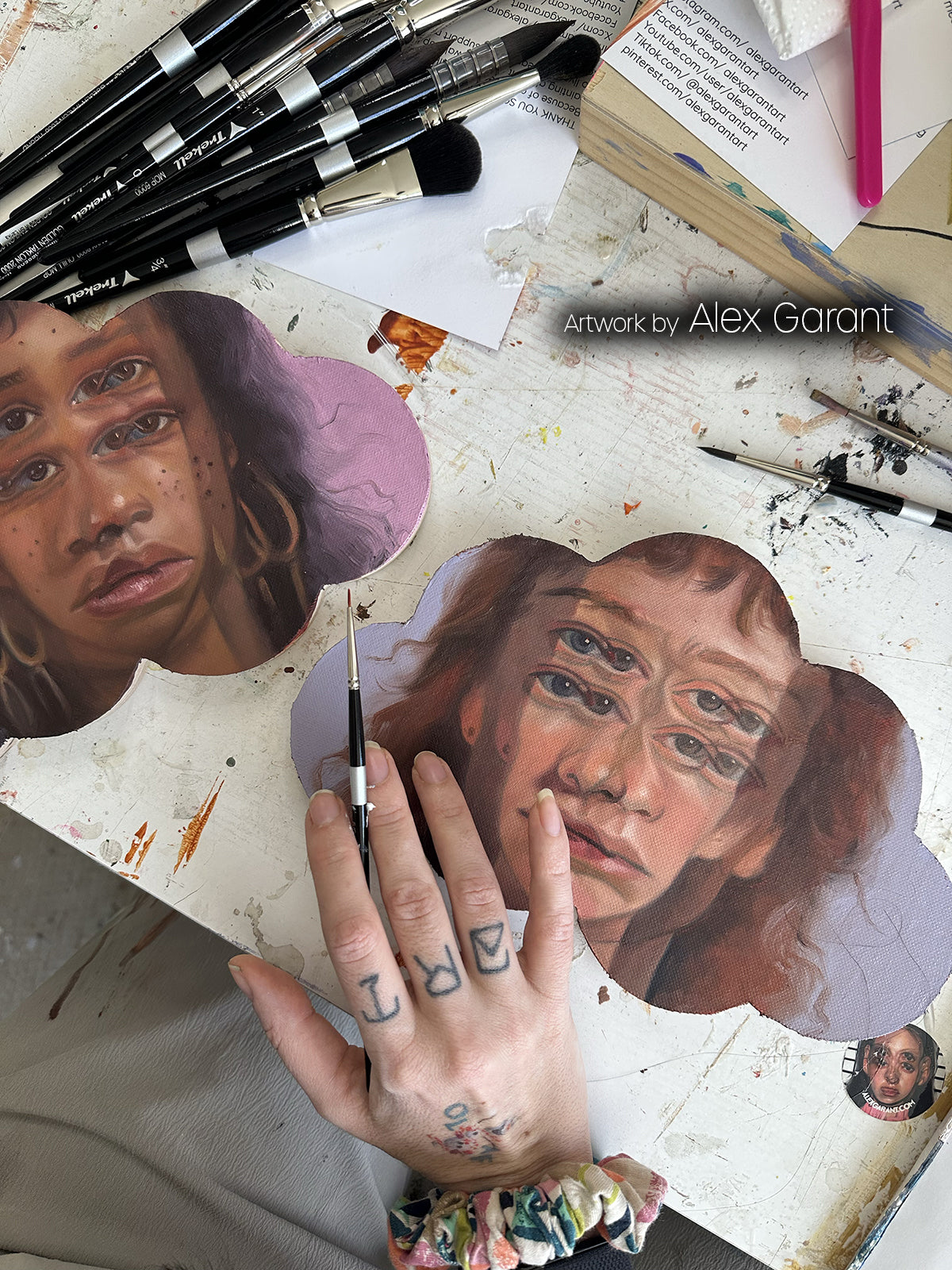 Two realistic painted faces on cloud-shaped panels beneath black-handled brushes, with an artist's hand in front.
