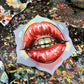 Glossy red lips with green teeth painted on a flower-shaped panel resting on a paint-splattered table.