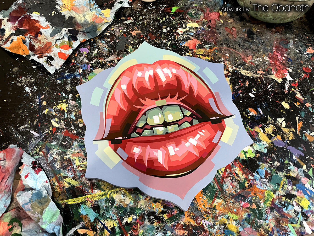 Glossy red lips with green teeth painted on a flower-shaped panel resting on a paint-splattered table.