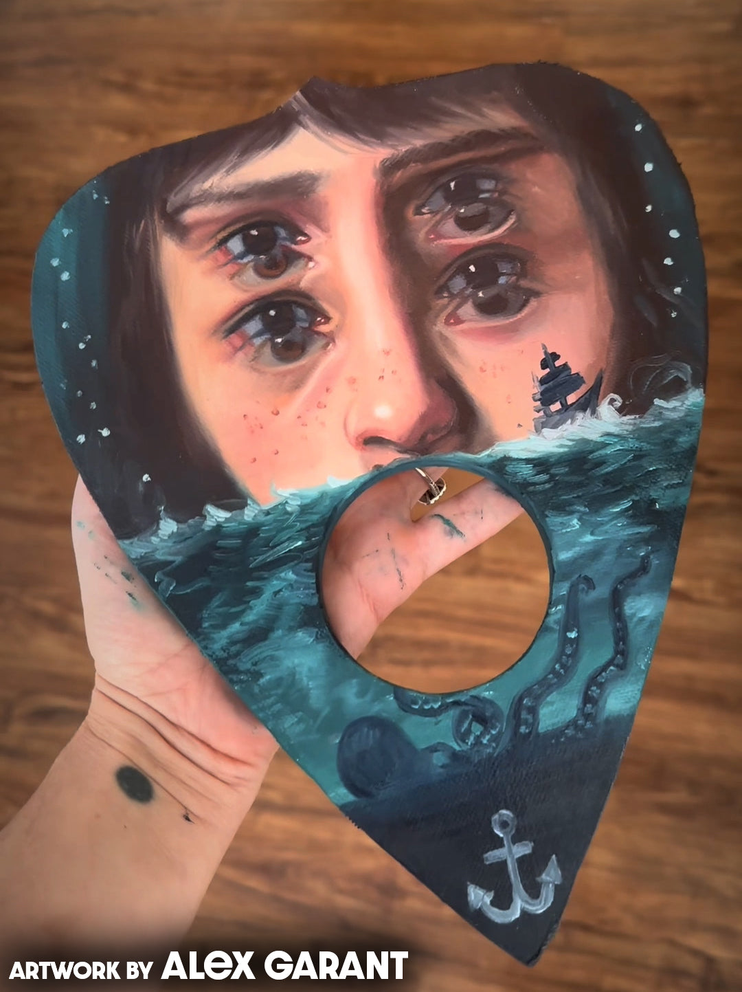 Four-eyed woman's face above teal ocean waves and an anchor on a planchette panel, credited to Alex Garant.