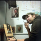 Artist in a cap holds a brush near his face while seated beside a small painting in a studio.