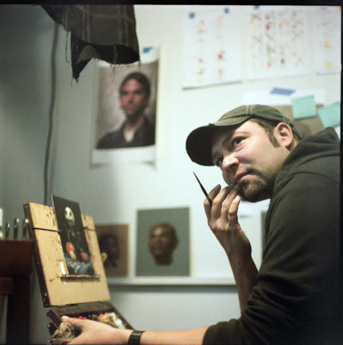 Artist in a cap holds a brush near his face while seated beside a small painting in a studio.