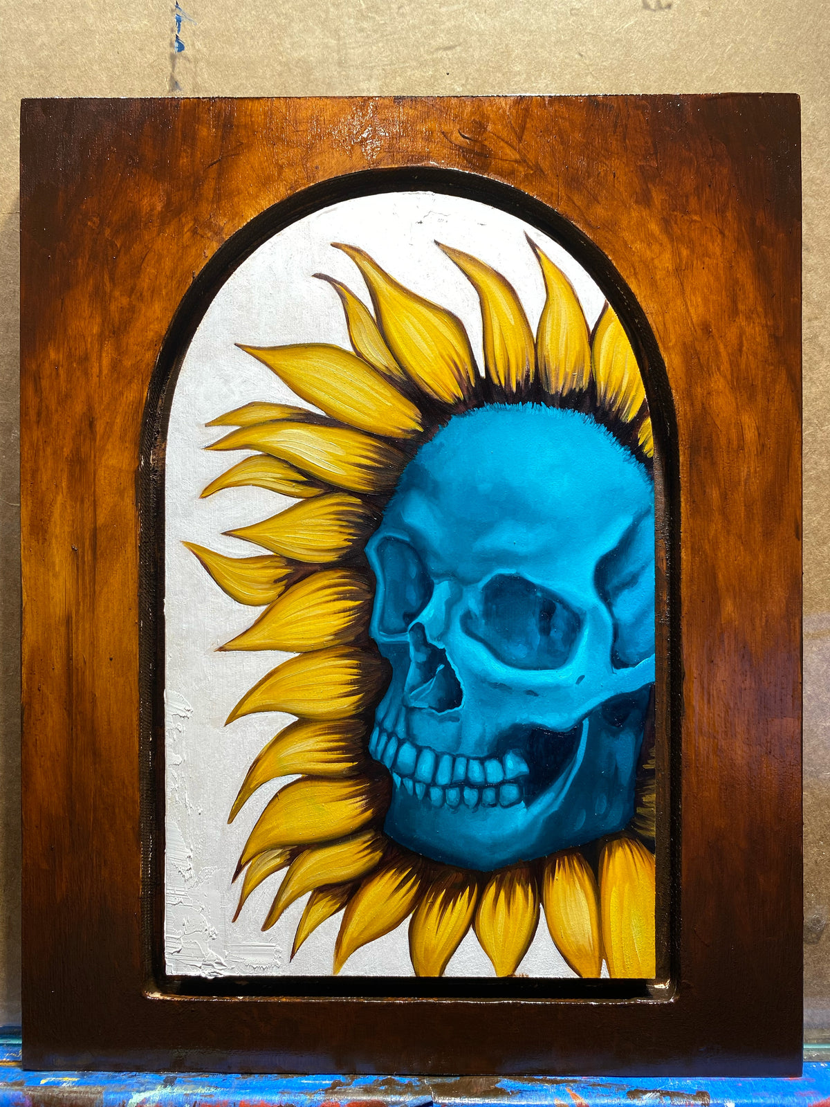 Blue skull surrounded by yellow sunflower petals inside an arched frame.