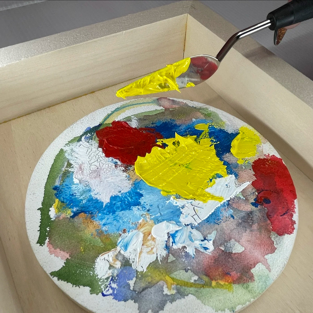 Palette knife lifts yellow paint from a round palette covered with blue, white, red and green paint.