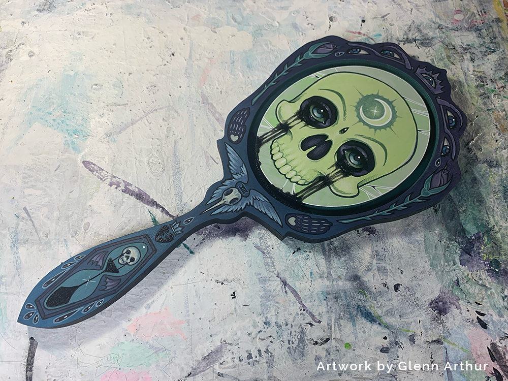 Green skull painted inside a dark ornate hand-mirror panel lying diagonally on a paint-marked surface.
