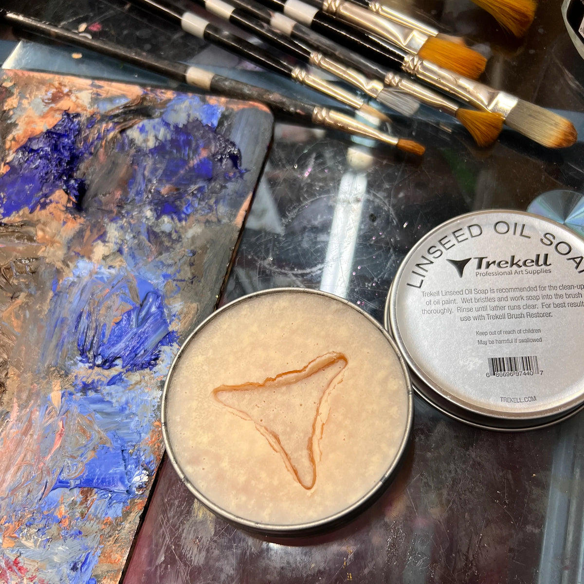 Open tin of pale linseed oil soap beside its lid, brushes and a paint-covered palette.