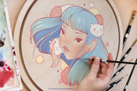 Hand paints a pastel blue-haired woman's portrait on a round panel beside brushes and paint containers.